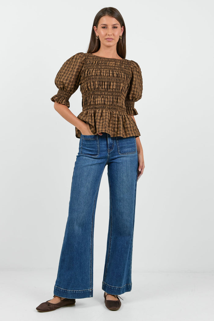 A woman wearing a chocolate and caramel checkered smocked top with puffed short sleeves, paired with high-waisted blue jeans.