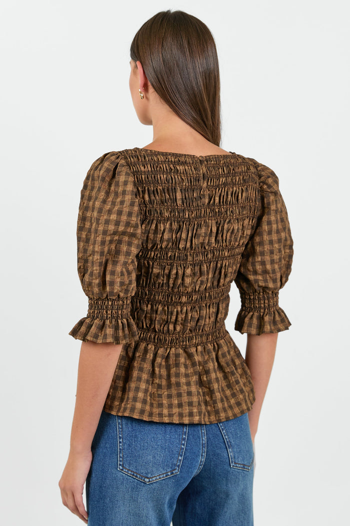 A woman wearing a chocolate and caramel checkered smocked top with puffed short sleeves, paired with high-waisted blue jeans.