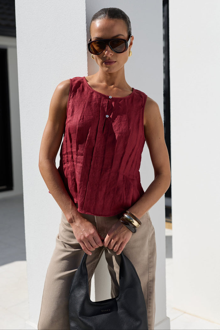 A woman modeling a sleeveless burgundy top with a buttoned neckline, paired with beige trousers and holding a black handbag, set against a modern white architectural background.