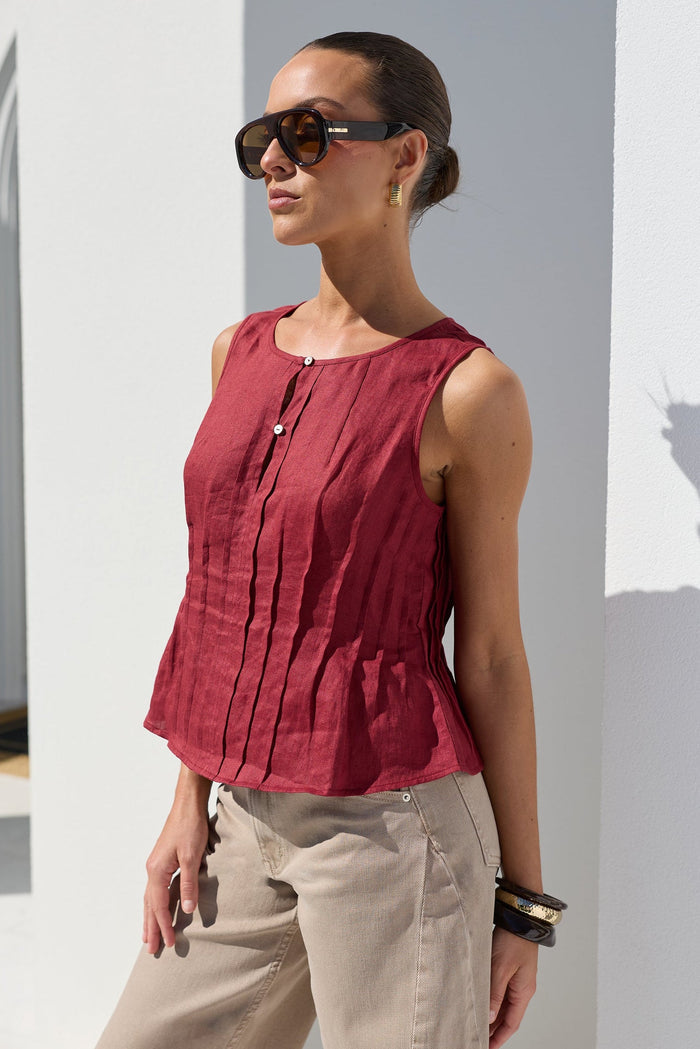 A woman wearing a sleeveless, deep red peplum top and beige trousers, standing in a bright white architectural setting with strong sunlight and shadows.