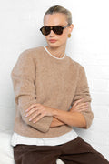 Woman wearing a beige sweater and sunglasses against a white brick wall.
