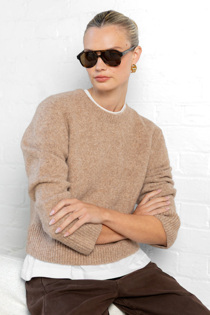 Woman wearing a beige sweater and sunglasses against a white brick wall.