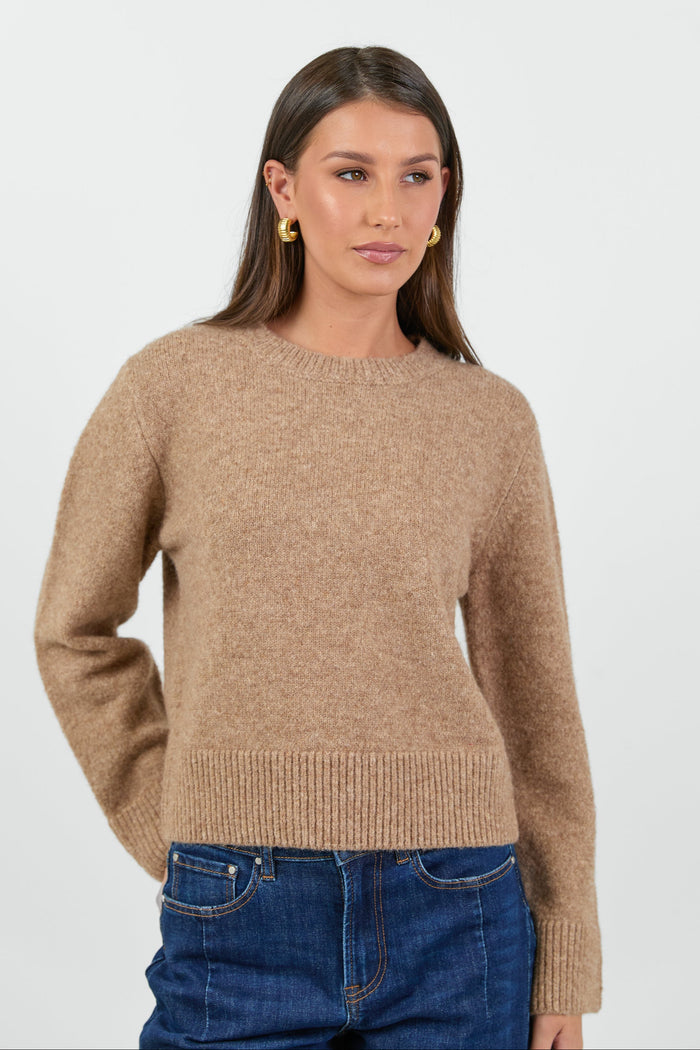 A woman wearing a soft, biscuit-colored crew neck jumper with long sleeves and ribbed cuffs, paired with dark blue high-waisted jeans.