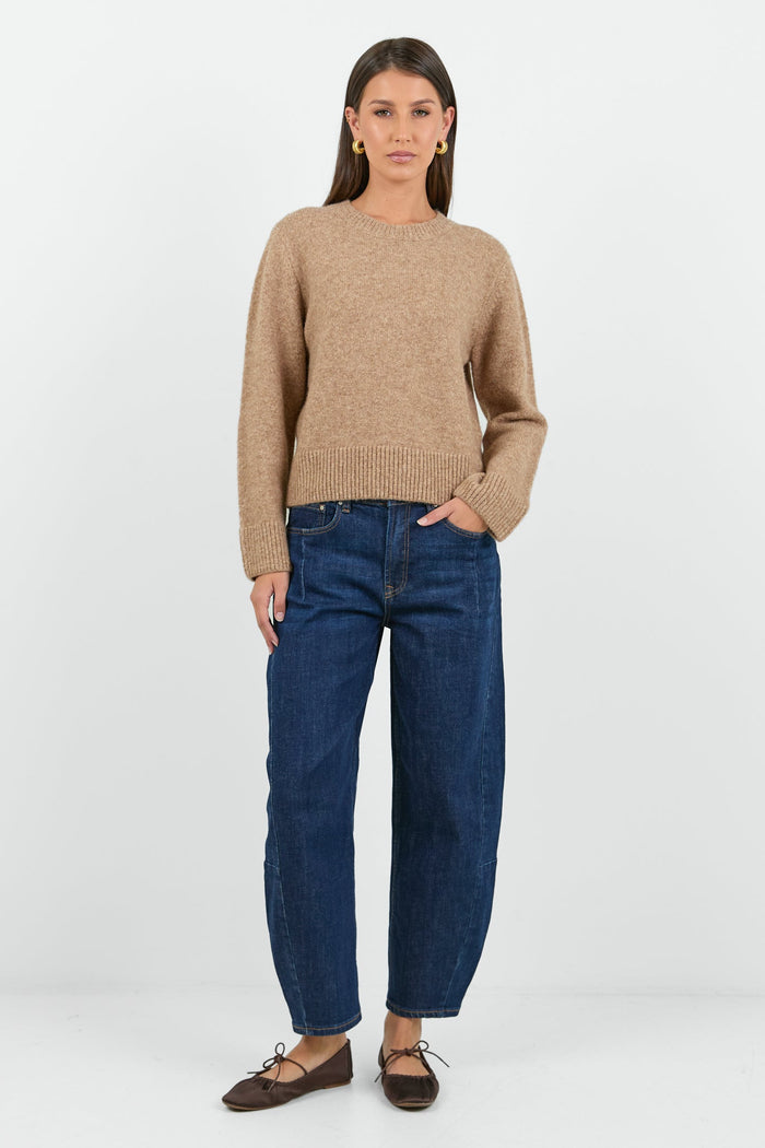 A woman wearing a soft, biscuit-colored crew neck jumper with long sleeves and ribbed cuffs, paired with dark blue high-waisted jeans.