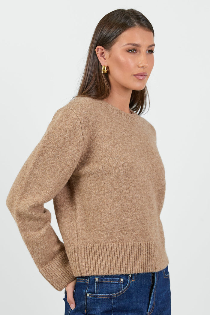 A woman wearing a soft, biscuit-colored crew neck jumper with long sleeves and ribbed cuffs, paired with dark blue high-waisted jeans.