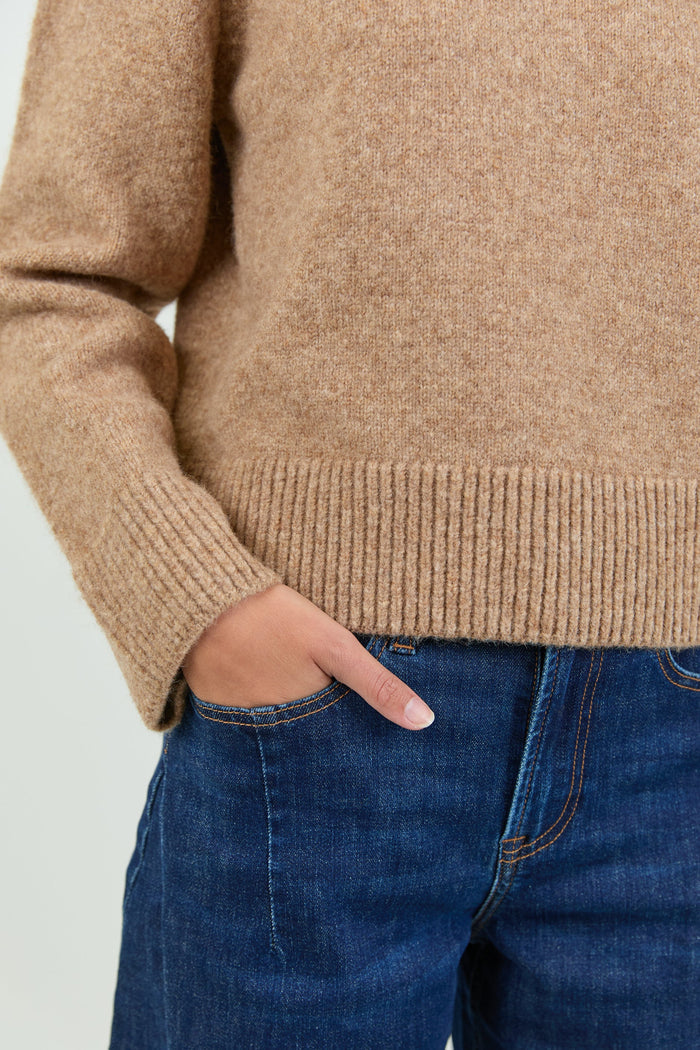A woman wearing a soft, biscuit-colored crew neck jumper with long sleeves and ribbed cuffs, paired with dark blue high-waisted jeans.