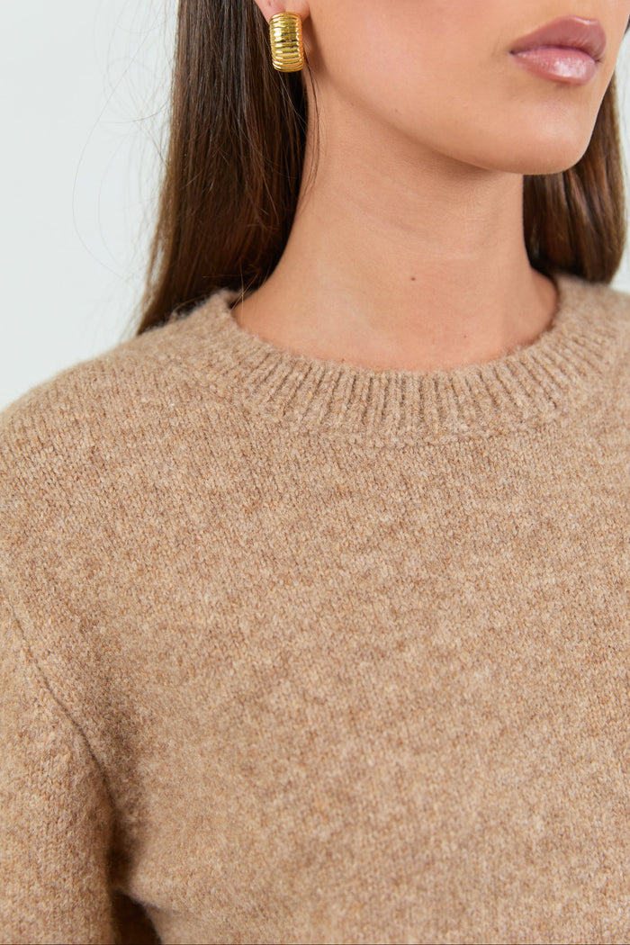 A woman wearing a soft, biscuit-colored crew neck jumper with long sleeves and ribbed cuffs, paired with dark blue high-waisted jeans.