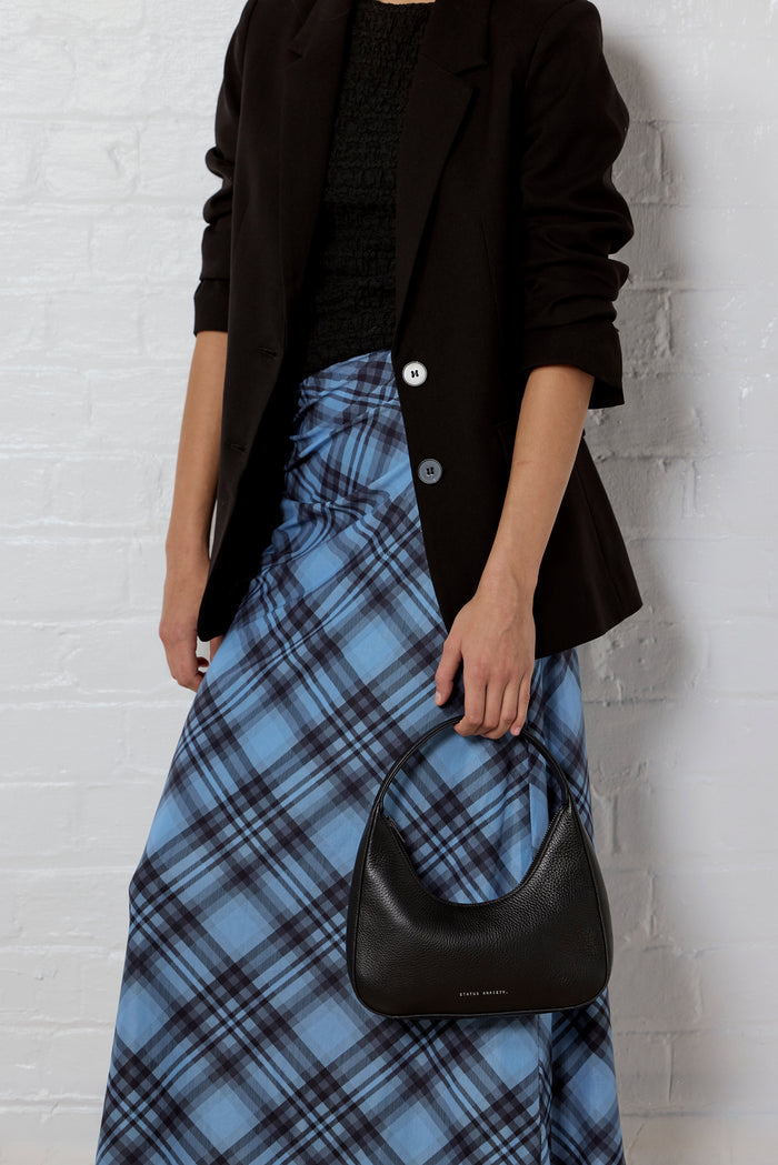 A model wearing a black blazer, black textured top, and a blue plaid skirt, holding a black crescent-shaped leather handbag against a white brick wall.