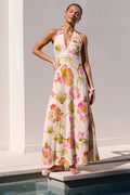 A woman models a sleeveless halter-neck maxi dress with a pink, green, and yellow floral watercolor print, standing on the edge of a swimming pool against a white stucco wall.