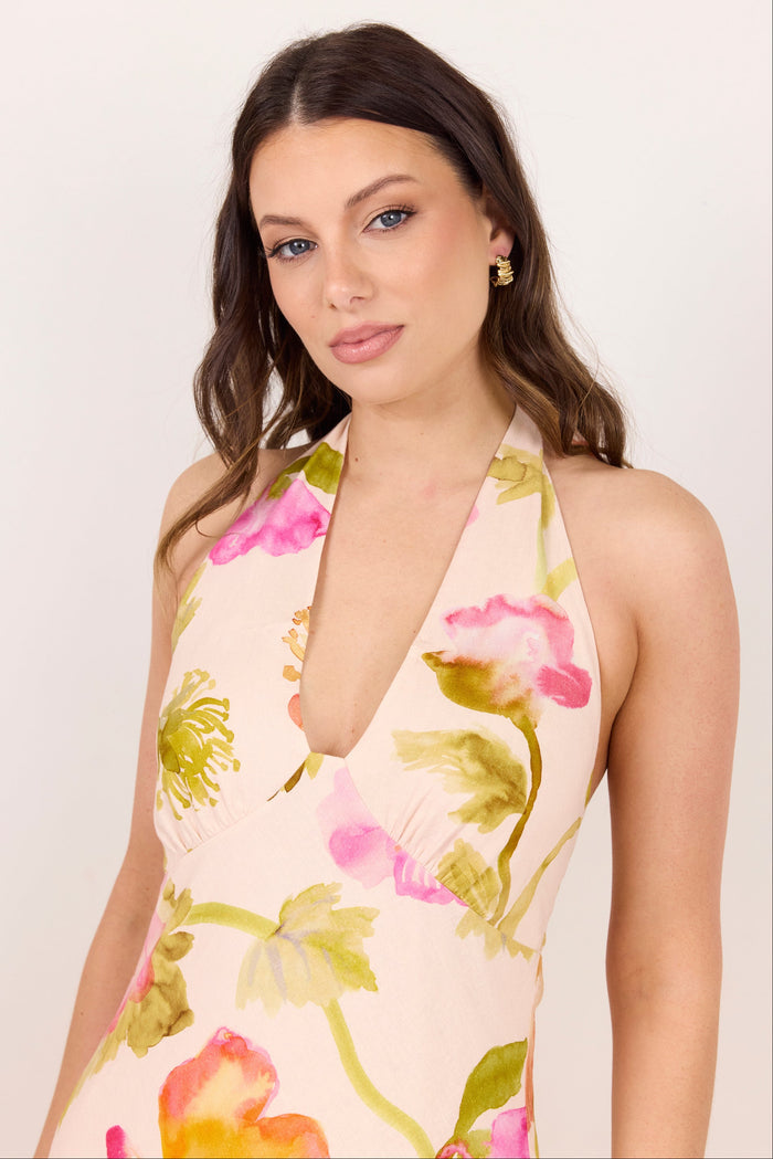A woman wearing a halter-neck dress with a vibrant pink, green, and orange floral print on a light beige background, photographed against a clean white studio backdrop.