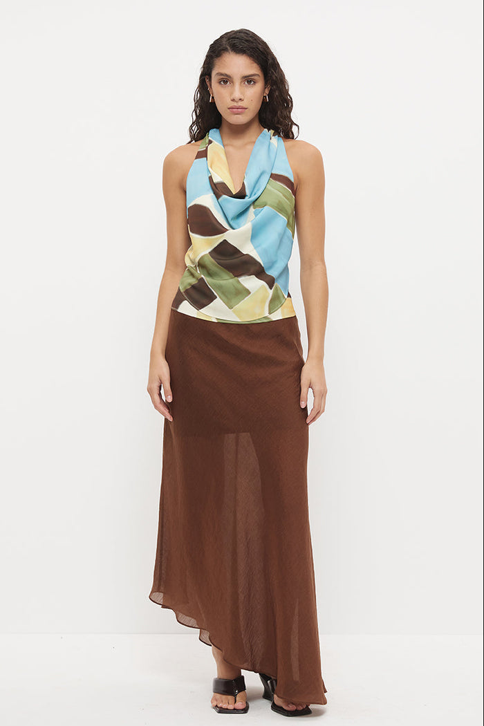 A model stands against a plain white studio background wearing a sleeveless halter-neck maxi dress with a multicolor abstract print on the bodice and a flowing brown skirt.