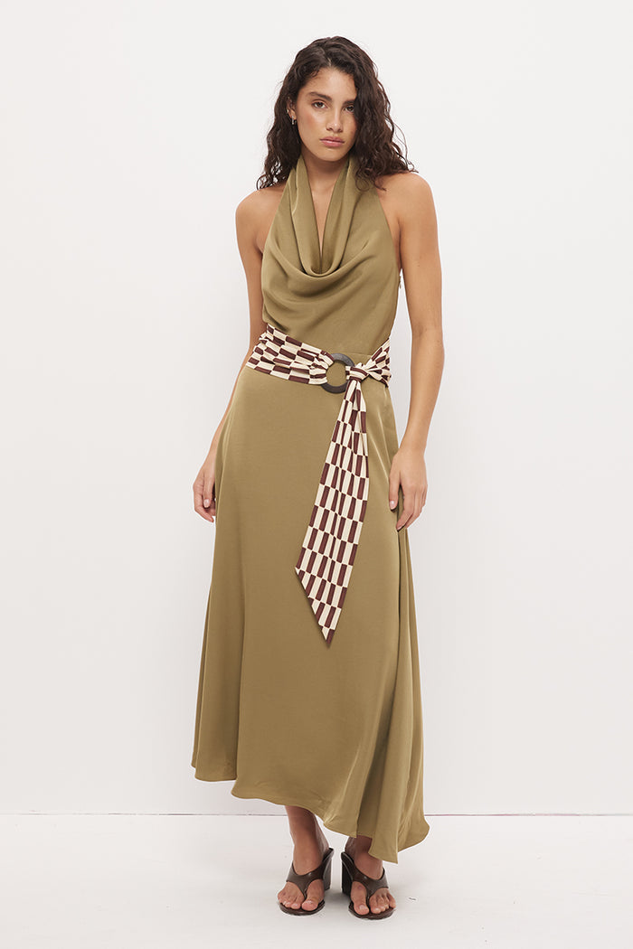 A woman modeling a sleeveless olive green halter maxi dress with a cowl neck, a matching tie belt, and a brown and white checkered patterned scarf tied at the waist, set against a plain white studio background.