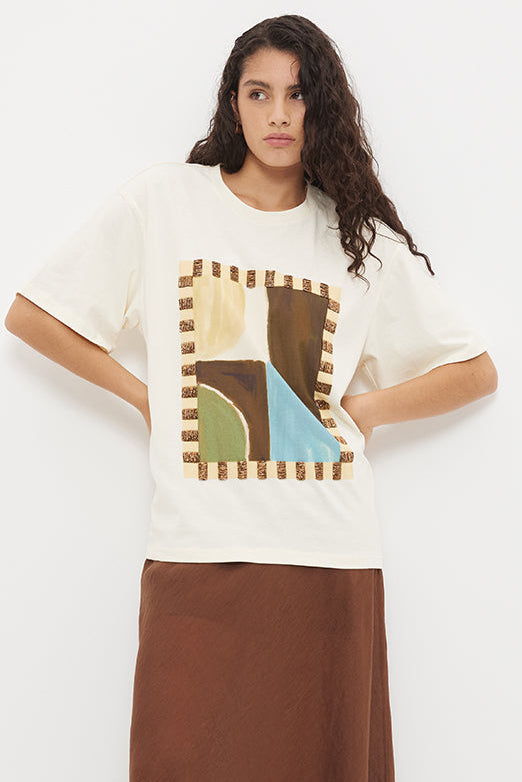 A woman modeling a cream-colored oversized t-shirt with a colorful geometric graphic print on the front, paired with a flowing brown maxi skirt and flat sandals, set against a plain white studio background.