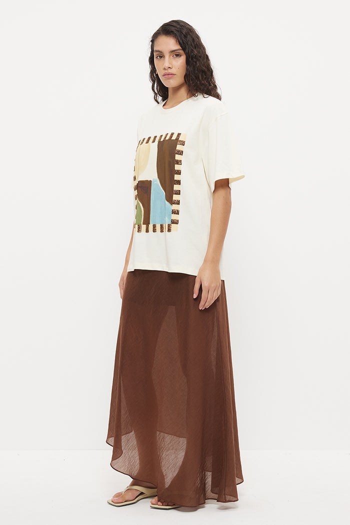 A woman modeling a cream-colored graphic t-shirt with a brown abstract print and a long brown sheer maxi skirt, standing against a plain white studio background.