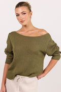 A woman wearing an olive green off-the-shoulder knit sweater and cream drawstring pants, photographed against a clean off-white studio background.