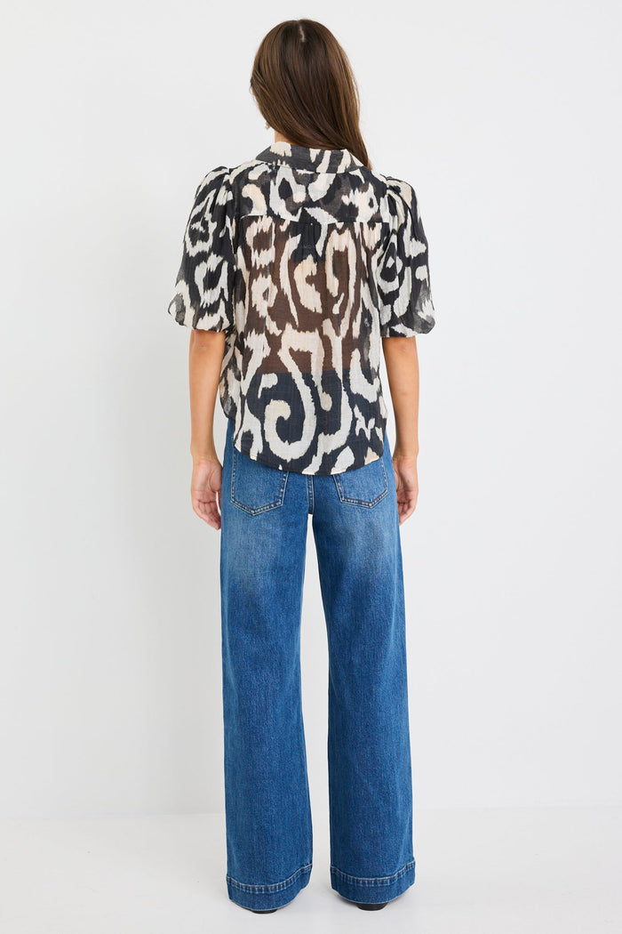 Woman wearing a black and cream zebra print short sleeve shirt with puff sleeves, button front, and tie waist detail, styled with blue jeans.