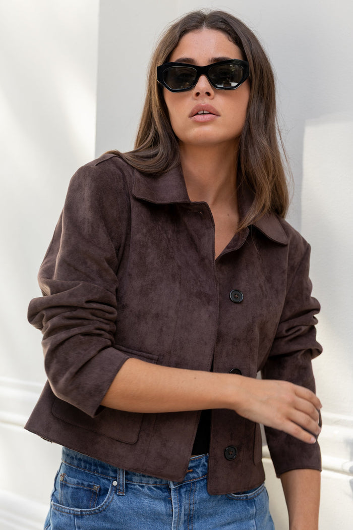 Woman wearing a brown jacket and blue jeans against a white wall.