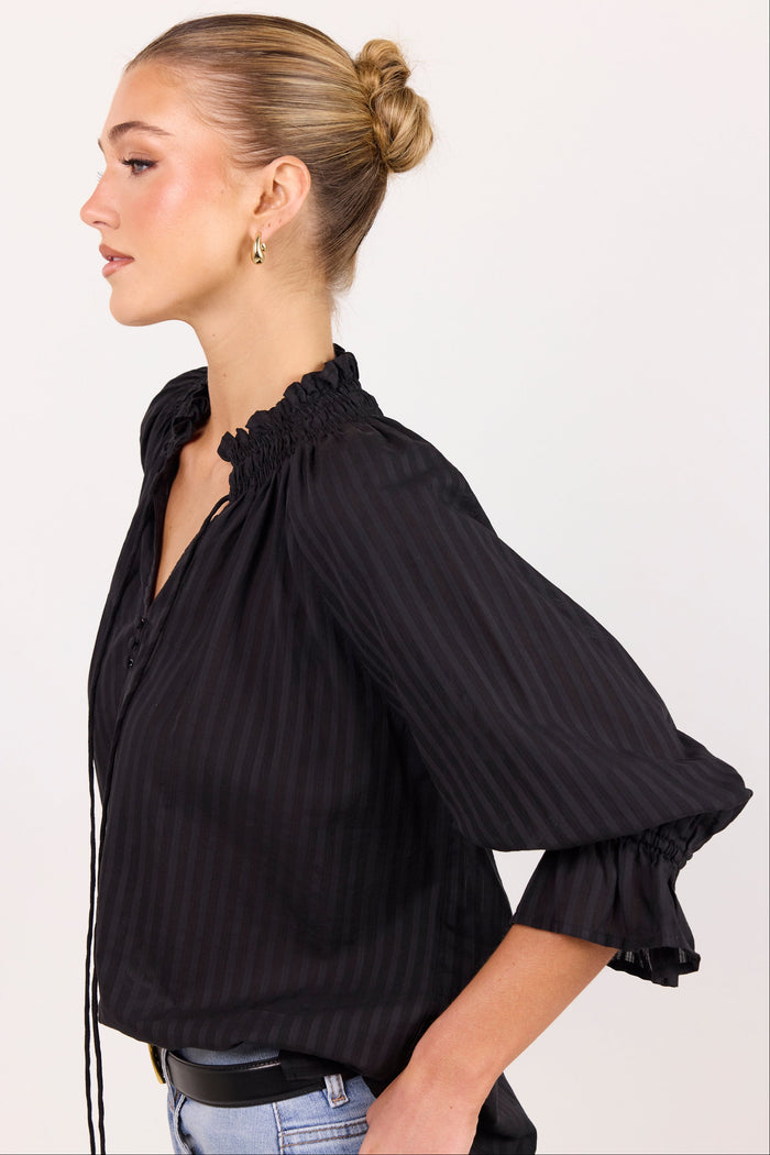 A woman shown in profile wearing a black pinstriped blouse with a smocked ruffled collar and three-quarter ruffled sleeves, paired with light blue jeans against a plain light background.