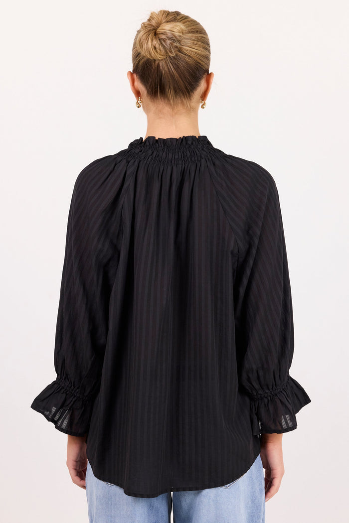 A woman viewed from the back wearing a black long-sleeve blouse with vertical pinstripes, a gathered smocked neckline, and ruffled cuffs, paired with light blue jeans against a plain white studio background.