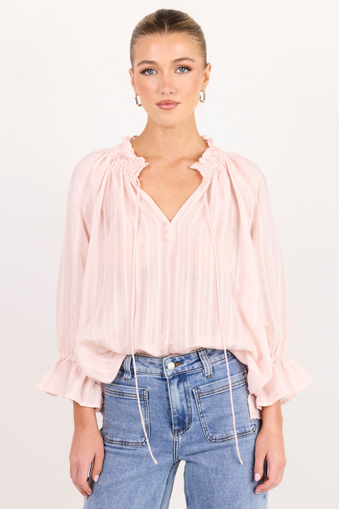 A woman modeling a light pink pleated blouse with a split neckline and ruffled cuffs, paired with light blue jeans, against a plain white studio background.