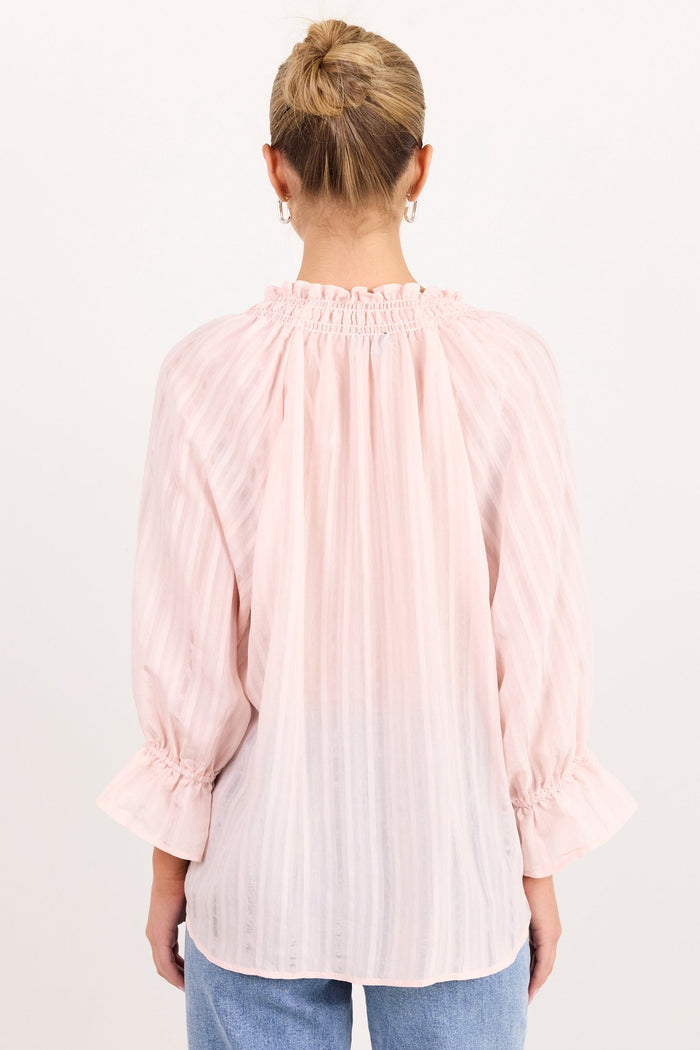 A woman viewed from behind wearing a light pink pleated blouse with ruffled cuffs and a smocked neckline, paired with light blue jeans, against a plain white background.
col_pattern