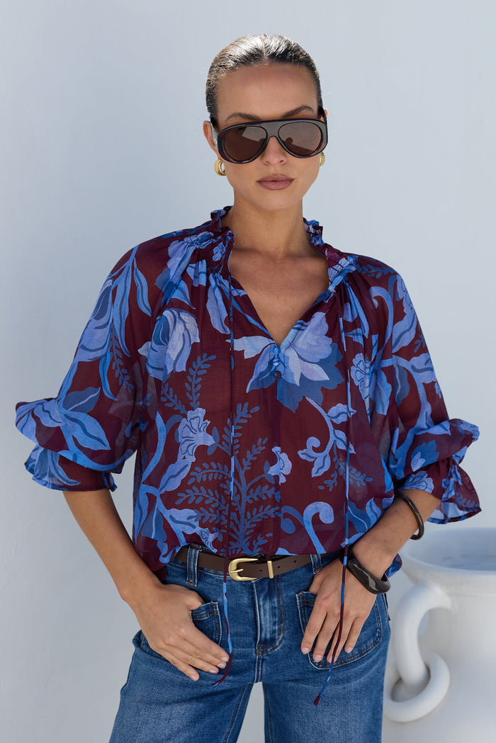 A woman modeling a sheer blue and burgundy floral print blouse with ruffled sleeves, paired with blue jeans and sunglasses, standing against a white wall.