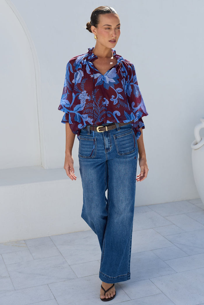 A woman stands outdoors against a white stucco wall wearing a blue and burgundy patterned blouse with ruffled sleeves, blue wide-leg jeans, and black sandals.