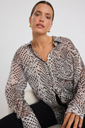 Woman wearing a loose-fitting, long-sleeve button-up shirt in a charcoal snake print, styled half-tucked into black jeans with gold hoop earrings.