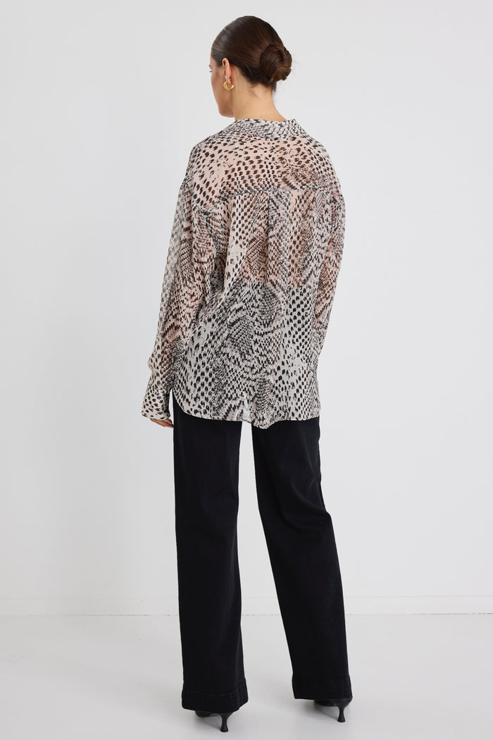 Woman wearing a loose-fitting, long-sleeve button-up shirt in a charcoal snake print, styled half-tucked into black jeans with gold hoop earrings.
