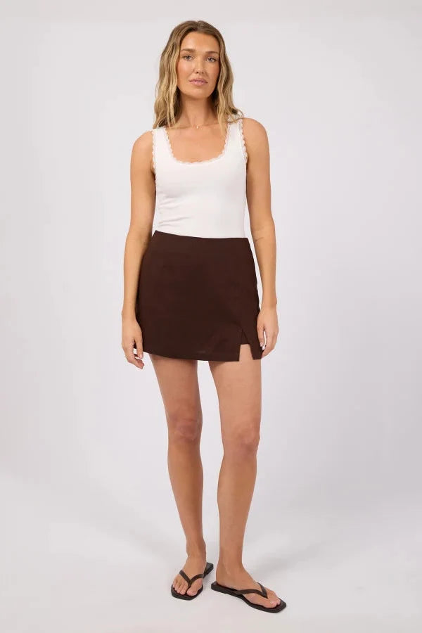 A woman modeling a white sleeveless top with scalloped edges and a dark brown mini skirt, paired with black flip-flop sandals, against a light gray studio background.