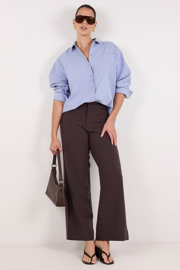 A model stands against a plain white studio background wearing a light blue button-down shirt, dark brown wide-leg trousers, black sandals, and holding a dark brown handbag.