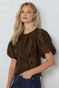 Woman wearing a brown blouse with puffed sleeves against a white brick wall.