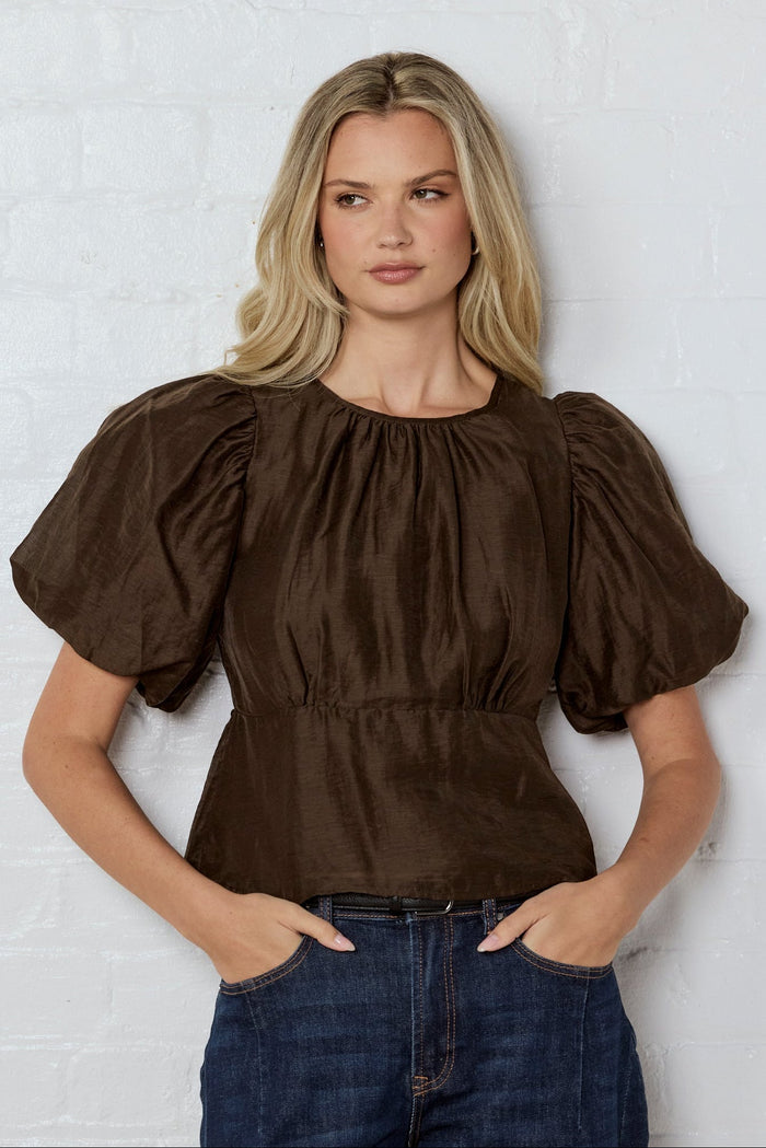 Woman wearing a brown blouse with puffed sleeves against a white wall