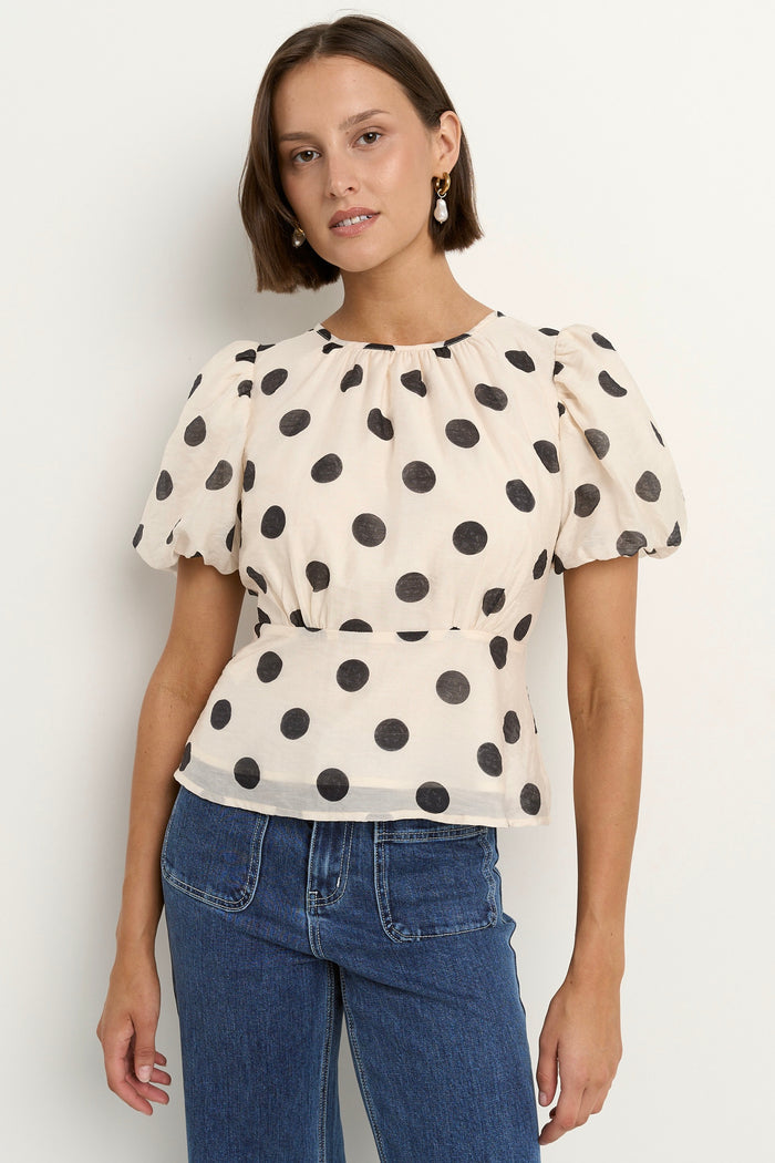 Woman wearing a cream short sleeve top with black polka dots, puff sleeves, and blue jeans.