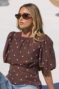 A woman wearing a brown short-sleeve top with white polka dots and denim shorts sits on the edge of a swimming pool, with two woven hanging chairs in the background.