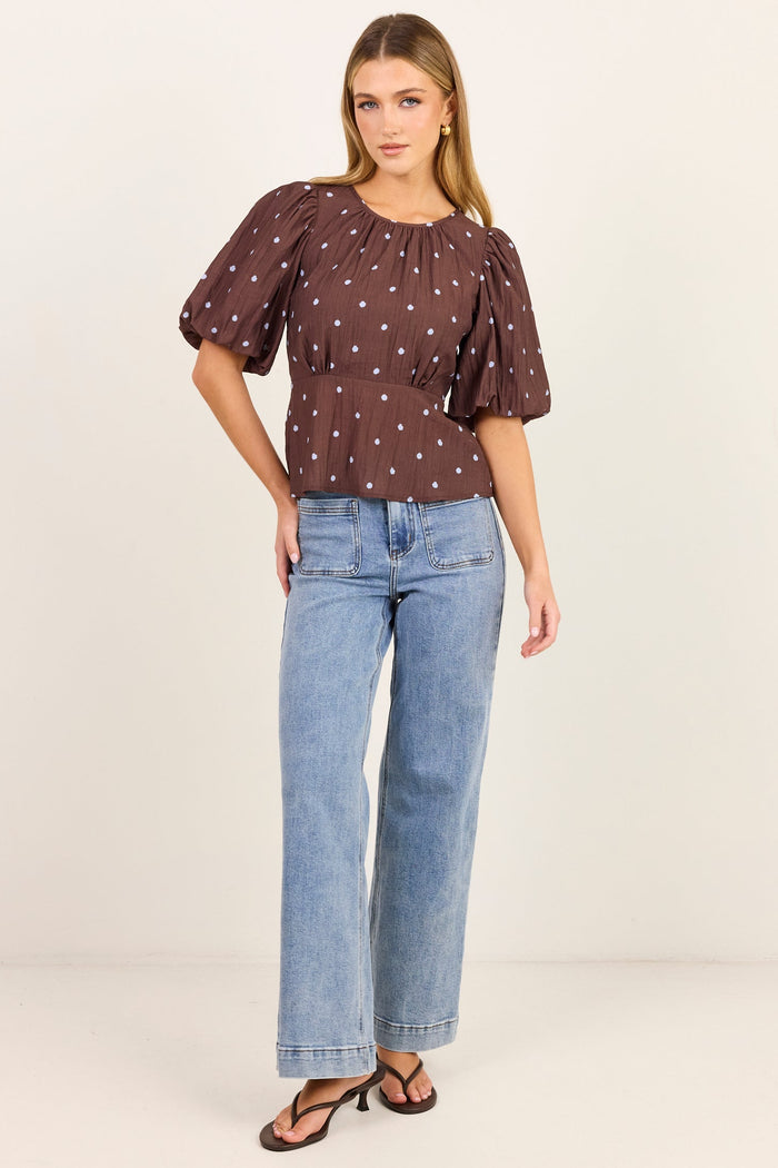 A woman modeling a brown short-sleeve blouse with white polka dots and puff sleeves, paired with light-wash blue jeans and brown sandals, against a plain off-white studio background.