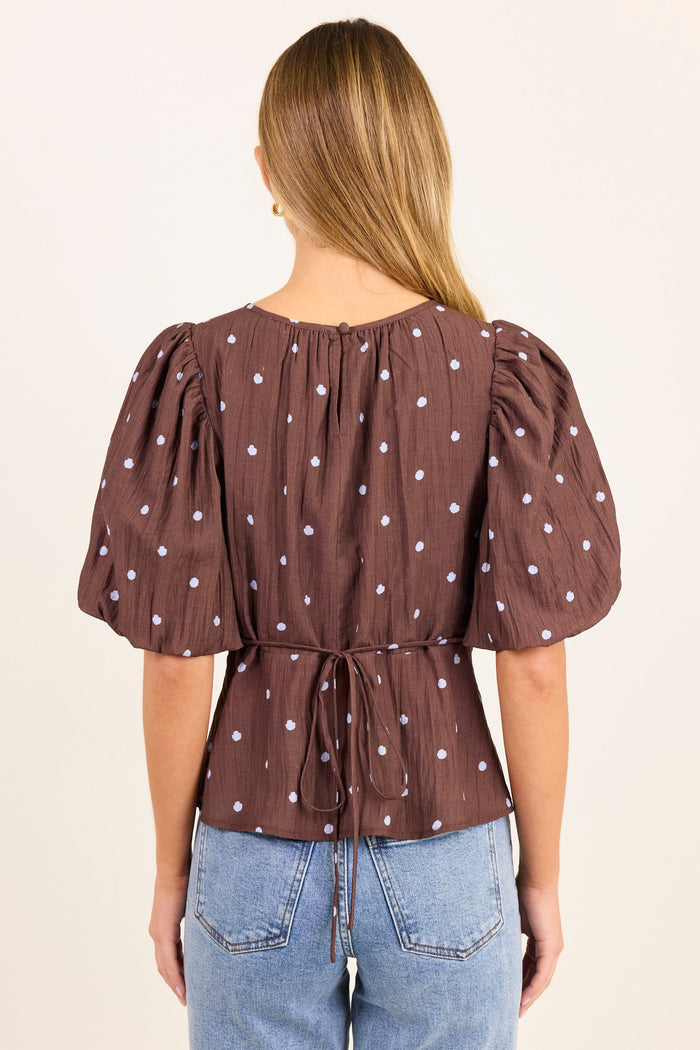 A woman viewed from the back wearing a brown short-sleeve top with white polka dots and puffed sleeves, paired with light blue jeans, against a plain cream background.