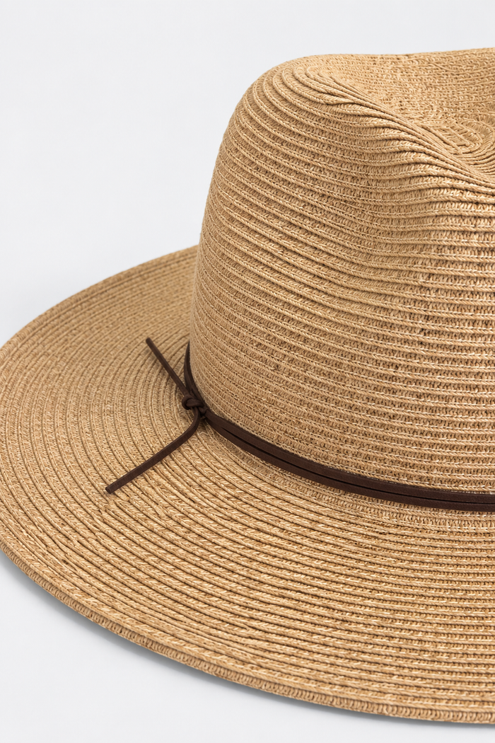A close-up of a woven straw fedora hat in a natural tan color, featuring a dark brown ribbon band tied in a bow on the side, set against a clean white background.