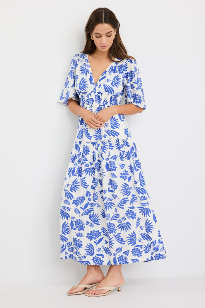 Woman wearing a white maxi dress with bold blue floral print, flutter sleeves, v-neckline, and tiered skirt, styled with neutral sandals.