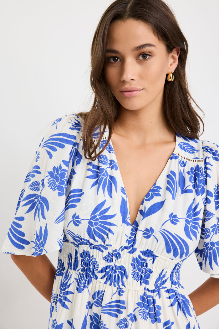 Woman wearing a white maxi dress with bold blue floral print, flutter sleeves, v-neckline, and tiered skirt, styled with neutral sandals.