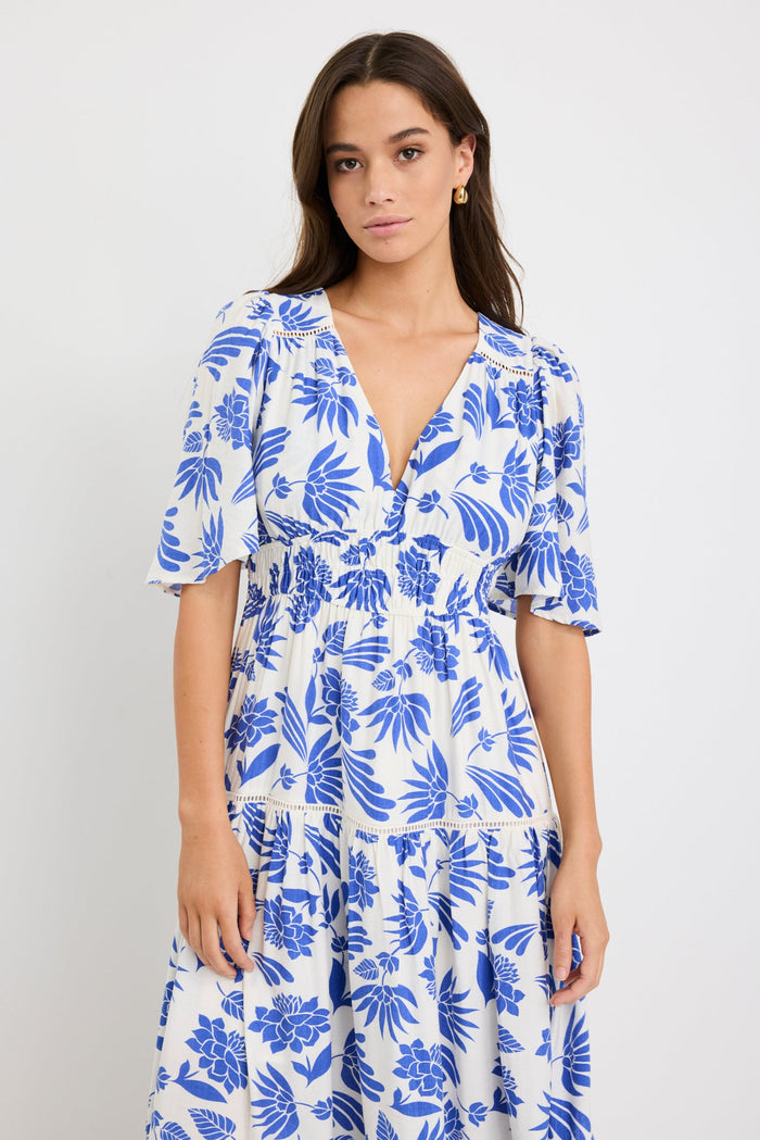Woman wearing a white maxi dress with bold blue floral print, flutter sleeves, v-neckline, and tiered skirt, styled with neutral sandals.