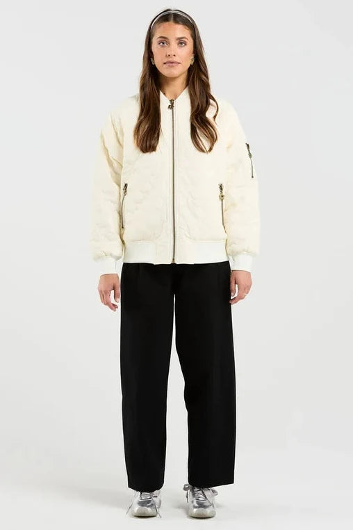 A woman stands against a plain light gray studio background wearing a cream zip-front jacket with black wide-leg pants and gray sneakers.