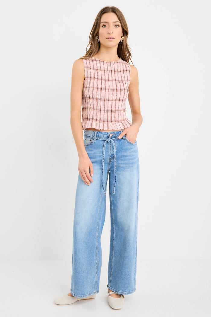 Woman wearing a blush pink sleeveless shirred top with check pattern and frilled hem, styled with light blue wide-leg jeans.