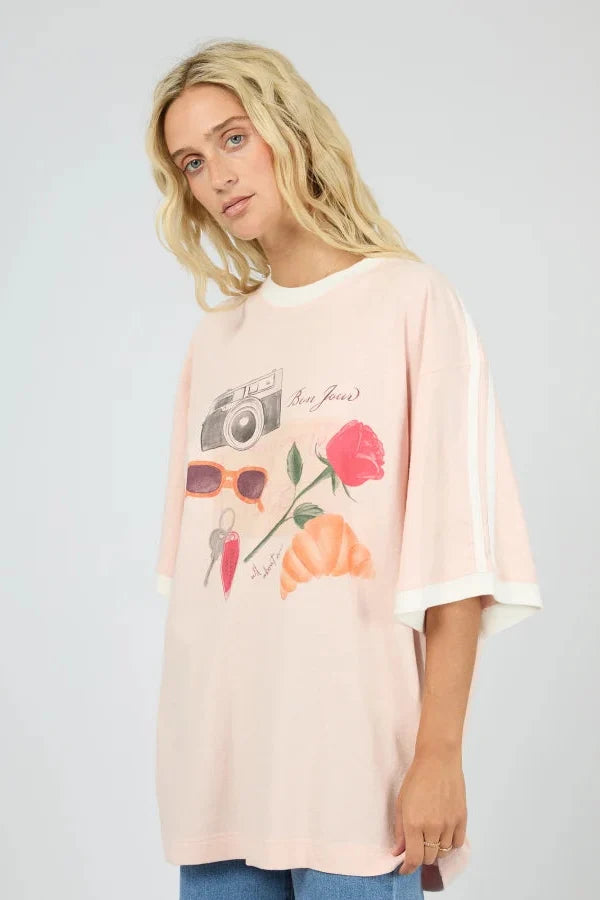 A woman wearing a light pink oversized graphic T-shirt with white sleeve stripes and a colorful front print featuring a camera, sunglasses, a rose, and a flower, paired with blue jeans against a light gray studio background.