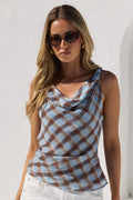 A woman modeling a sleeveless cowl-neck top with a brown and light blue plaid pattern, paired with white pants, standing against a white stucco wall with an archway.