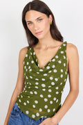 A woman wearing a sleeveless olive green cowl-neck top with a white polka dot pattern, paired with blue jeans, against a plain light gray background.