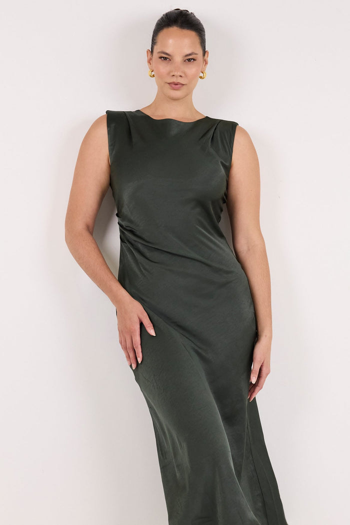 A woman modeling a sleeveless olive green midi dress with a draped, gathered design, paired with black strappy heels, against a plain white studio background.