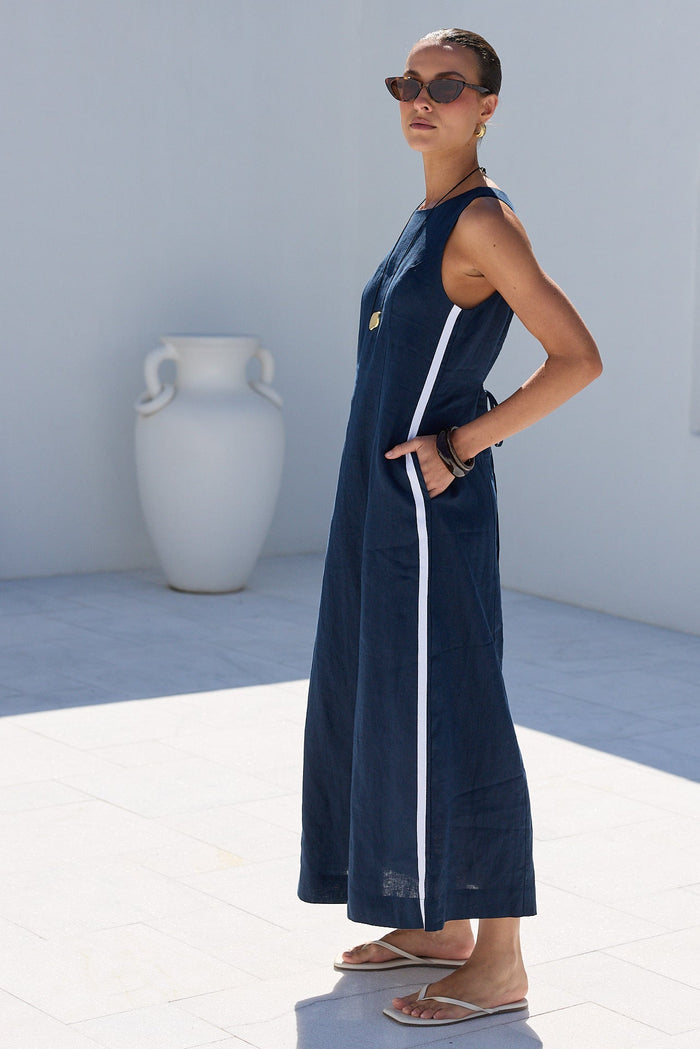 A woman wearing a sleeveless navy blue maxi dress with a white side stripe and sunglasses, standing in a bright white outdoor courtyard with a large white vase and dark green trees in the background.