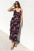 A woman wearing a black ruffled maxi dress with a pink and red floral print, standing against a white stucco wall next to a pool.