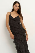 Woman wearing a black bias-cut polka dot dress with delicate straps and cascading ruffles, standing against a neutral wall.